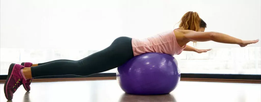 Person balancing face down on a purple exercise ball in a gym, arms and legs extended.