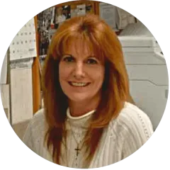 Smiling woman with long reddish hair in a white sweater, standing indoors in an office setting