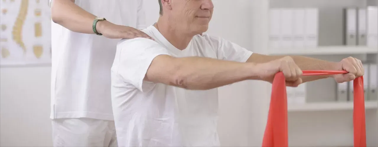 Person in white shirt exercises with a red resistance band while a therapist steadies their shoulder.