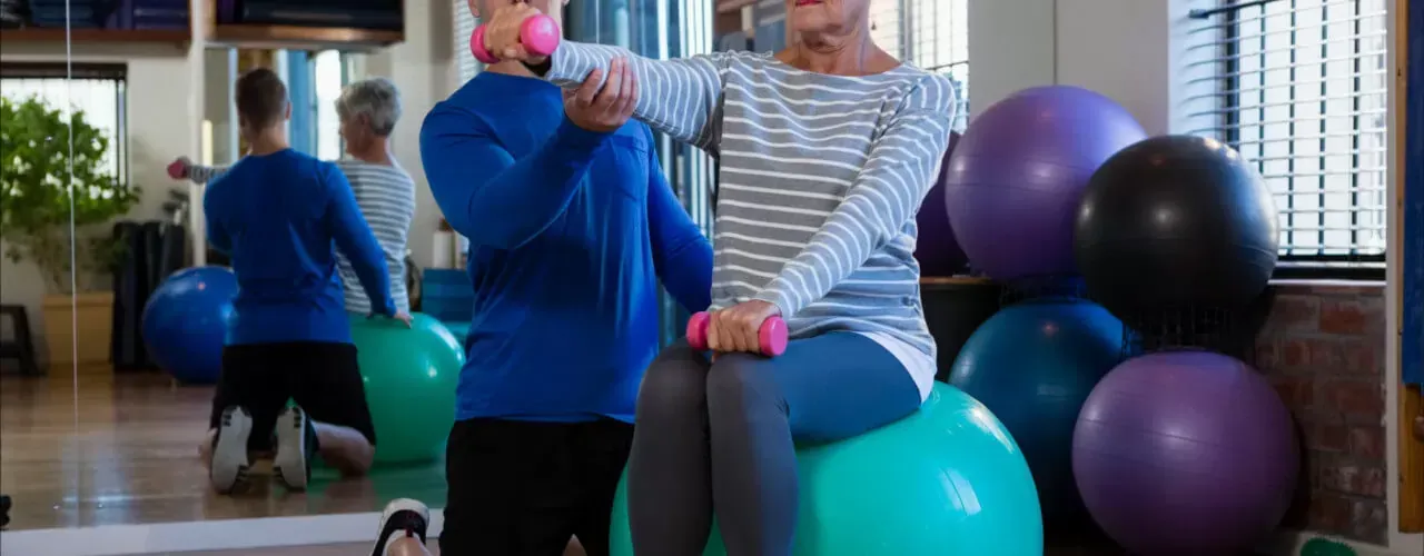 Two people exercising with stability balls in a bright gym, with mirrored walls and exercise equipment nearby