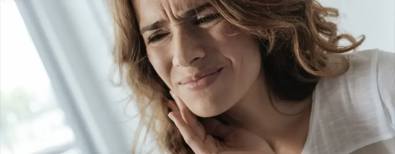 Woman grimacing and holding her jaw in pain indoors near a bright window