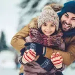 Smiling couple in winter coats and knit hats, hugging outdoors in a snowy setting