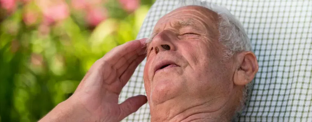 Elderly man resting outdoors, eyes closed, hand to forehead, with a pink flower background