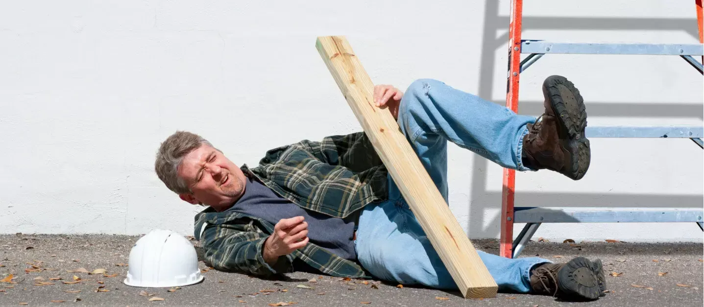 Worker lies on floor beside white hard hat, pinned under a wooden board near a ladder.