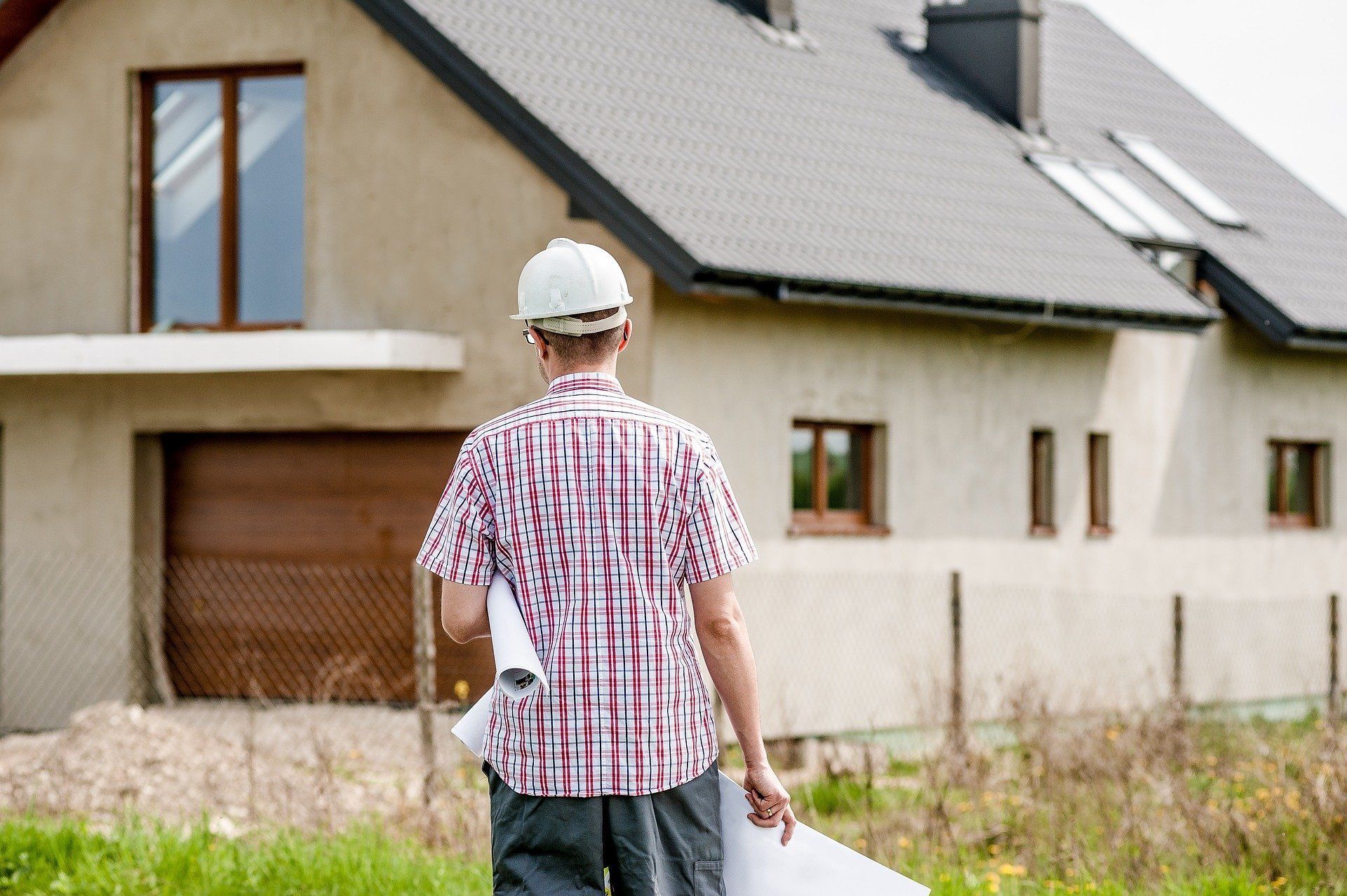 Ein Mann mit Bauhelm hält einen Bauplan vor einem im Bau befindlichen Haus.