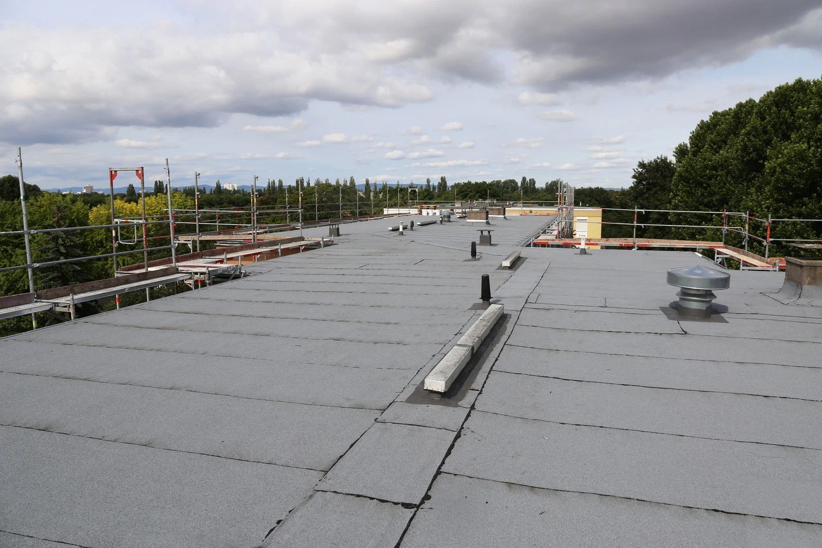 Flat rooftop with gray membrane, vents, and a cloudy sky over trees and railings.