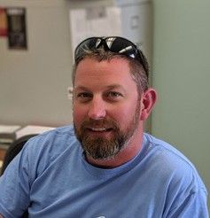 Man with beard wearing sunglasses on head, blue shirt, smiling indoors.