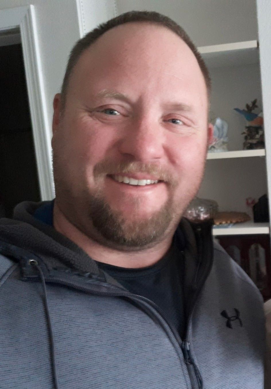 Man with beard, smiling, wearing gray hooded sweatshirt; indoor setting with white shelving visible.