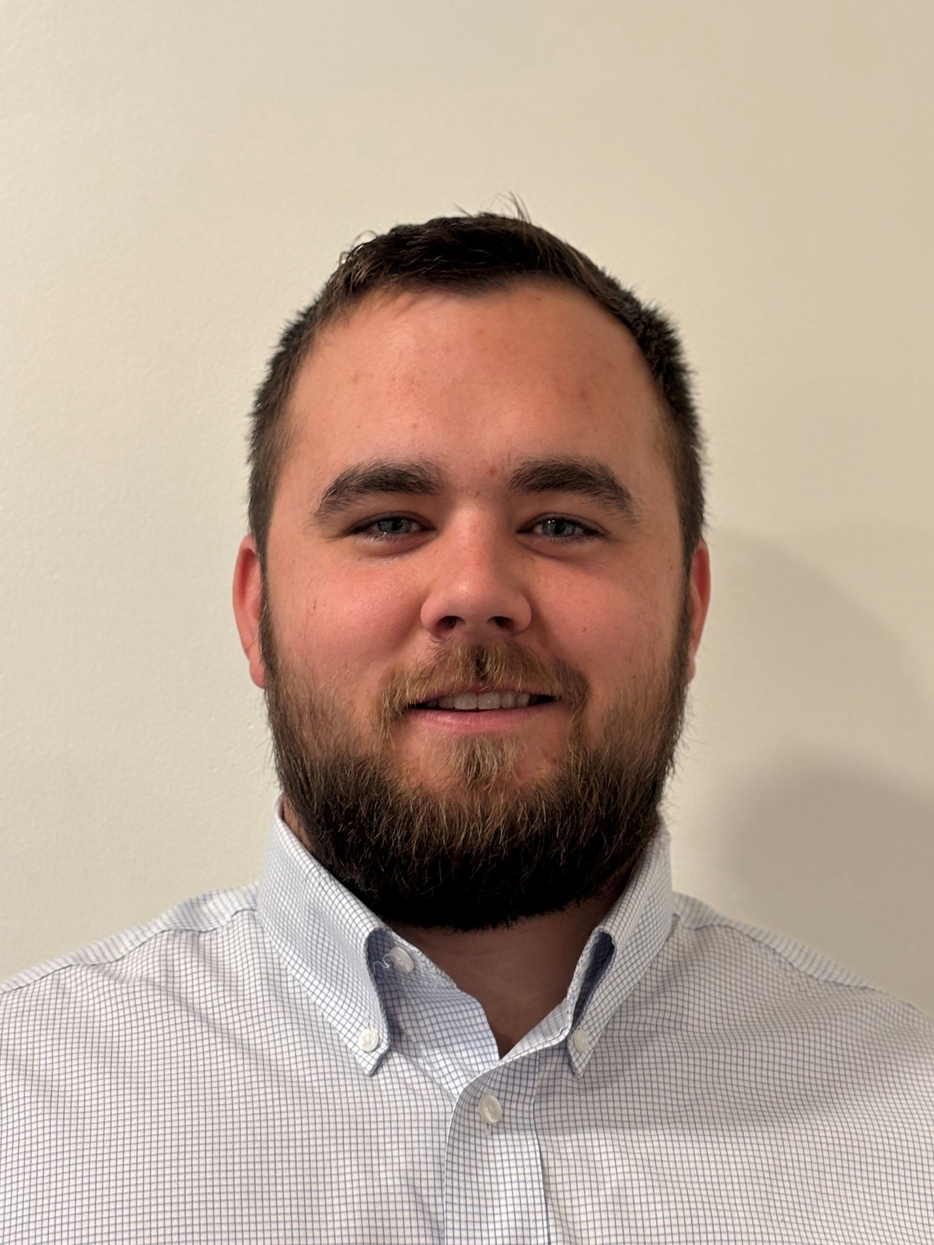 Man with a beard wearing a light blue and white patterned button-down shirt, smiles at the camera.
