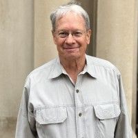 Man with gray hair and glasses smiles outdoors, wearing a light gray collared shirt.