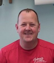 Man in red shirt smiling. Shirt has logo, possibly Falmouth Farm. Background is light blue.