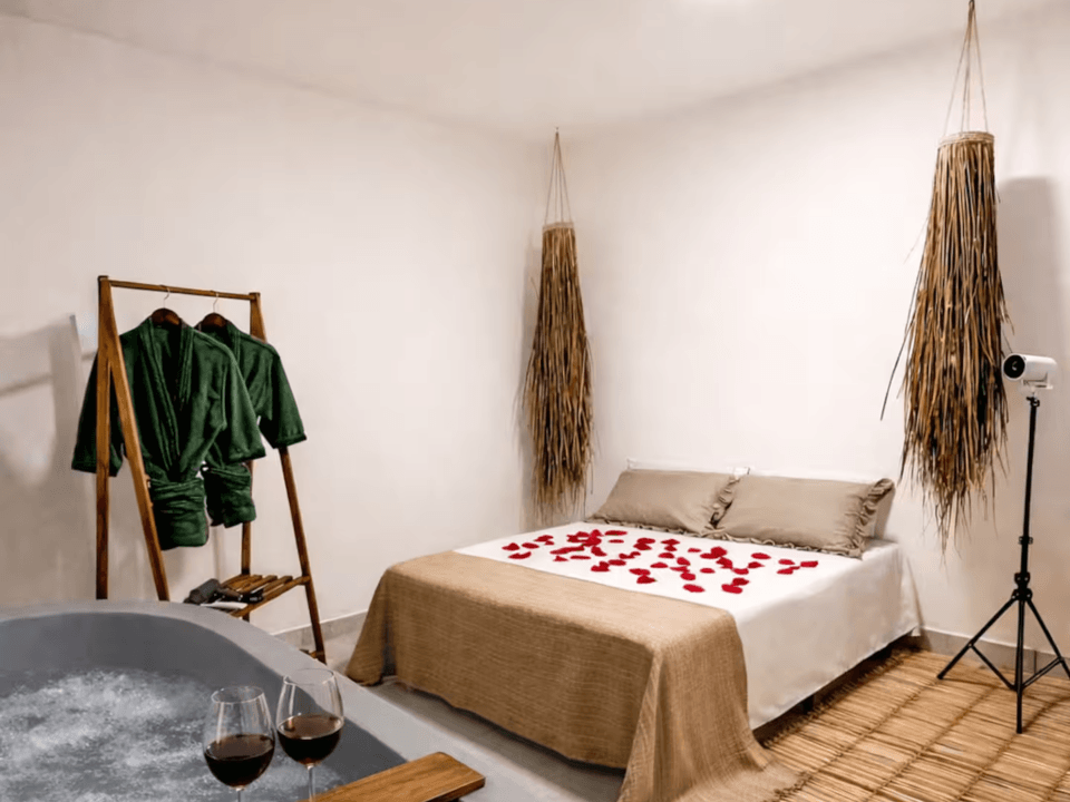 Quarto minimalista com cama, plantas secas penduradas, cabideiro e taças de vinho sobre uma mesa.