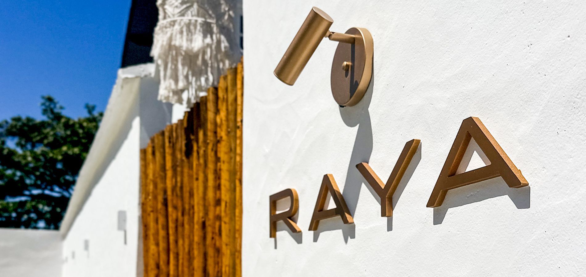 Detalhe da placa RAYA em uma parede branca com um toque decorativo dourado e céu azul.