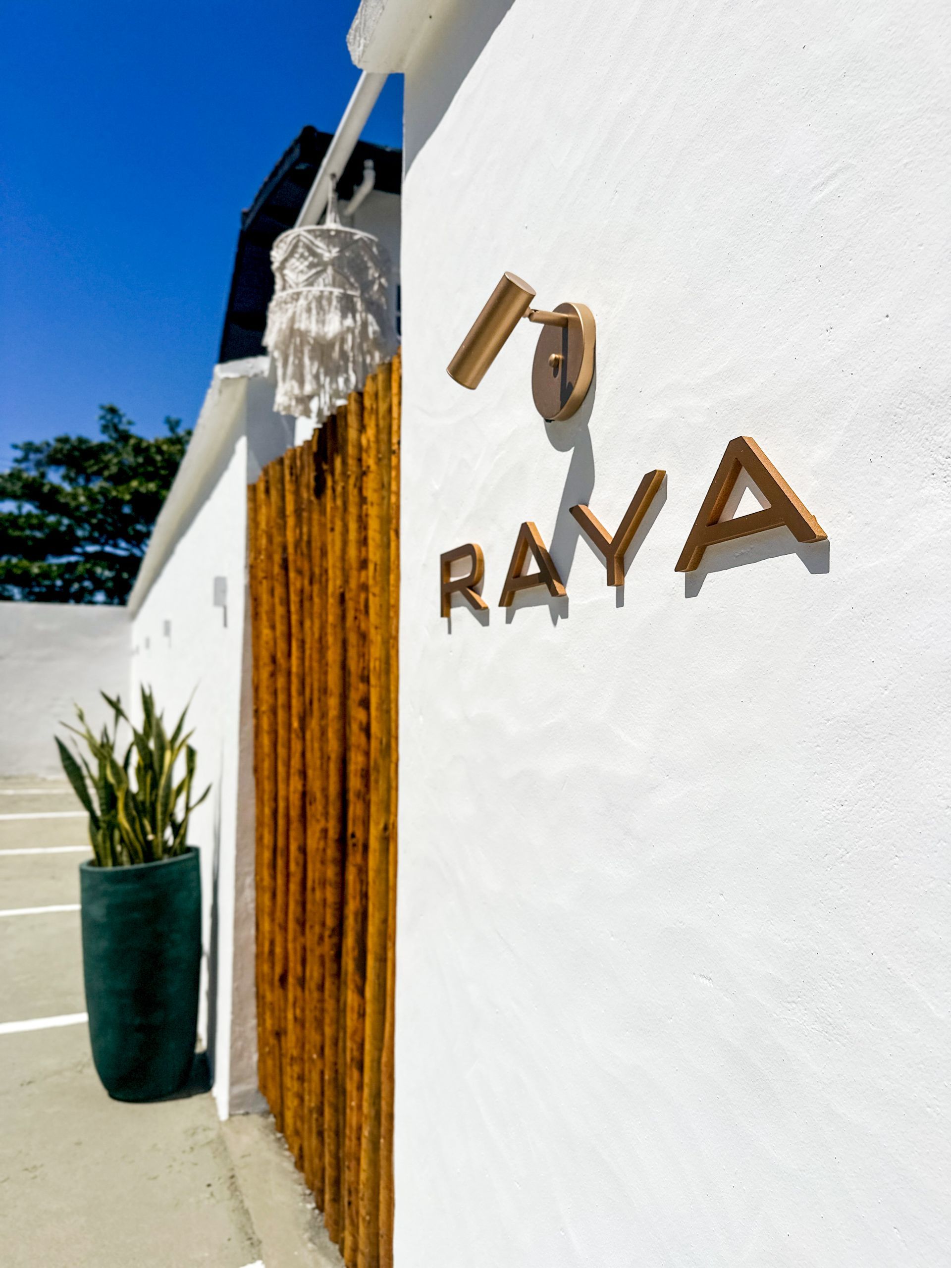 Placa RAYA em uma parede branca ao lado de uma porta de madeira e um vaso de planta sob a luz do sol.