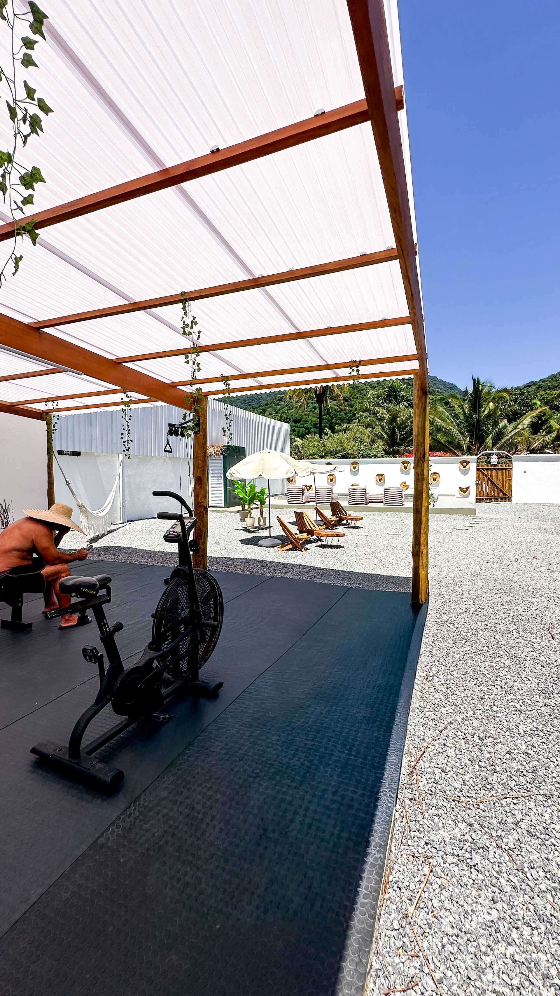 Área de ginástica ao ar livre com uma pessoa se exercitando em um remo ergométrico sob uma pérgola de madeira.
