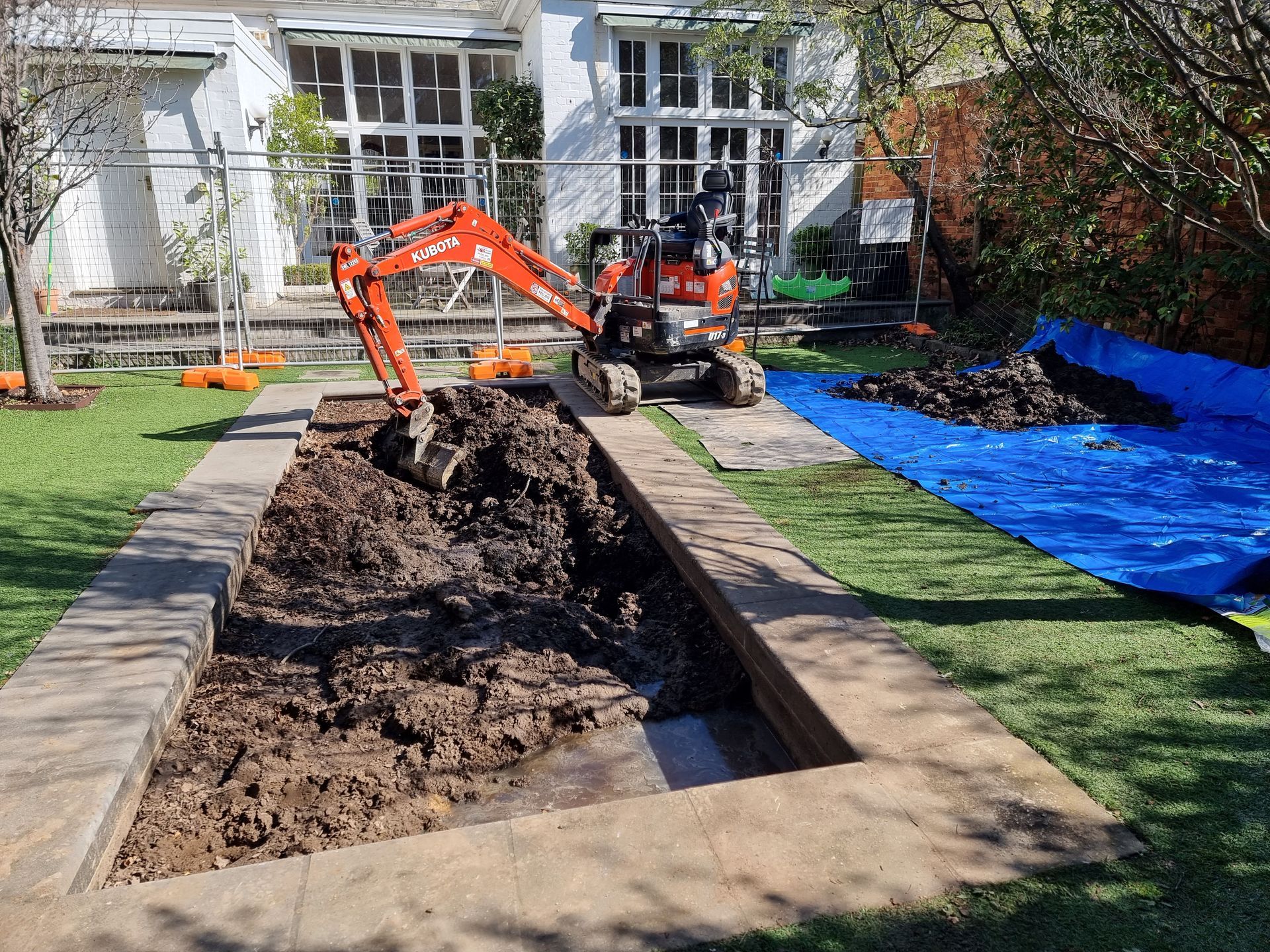 An Orange Excavator Digs Into a Rectangular Trench in a Grassy Backyard — All Over Pool Removals in Surrey Hills, VIC