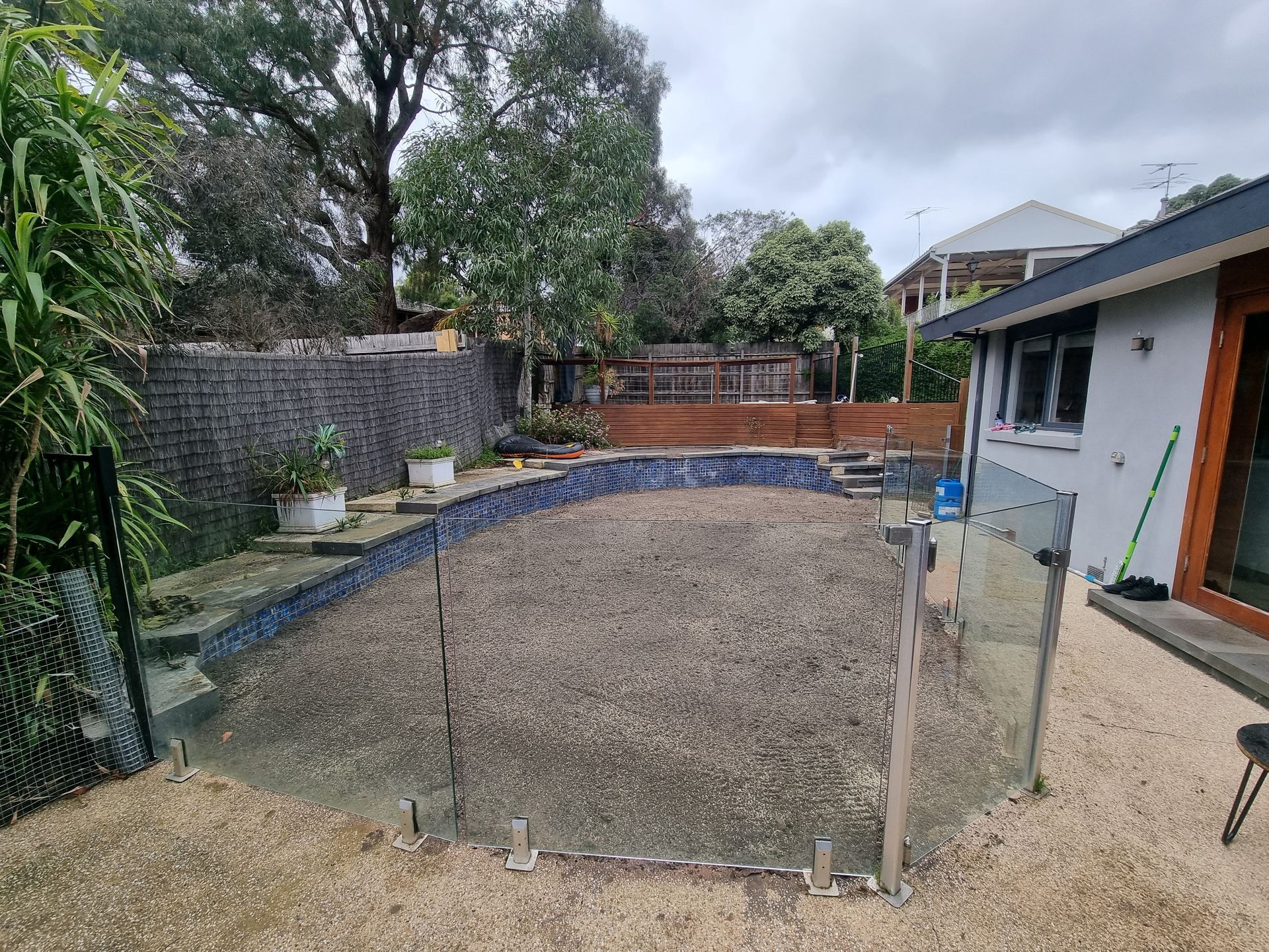 An Empty, Gravel-filled Swimming Pool Surrounded by a Glass Fence — All Over Pool Removals in Doncaster, VIC