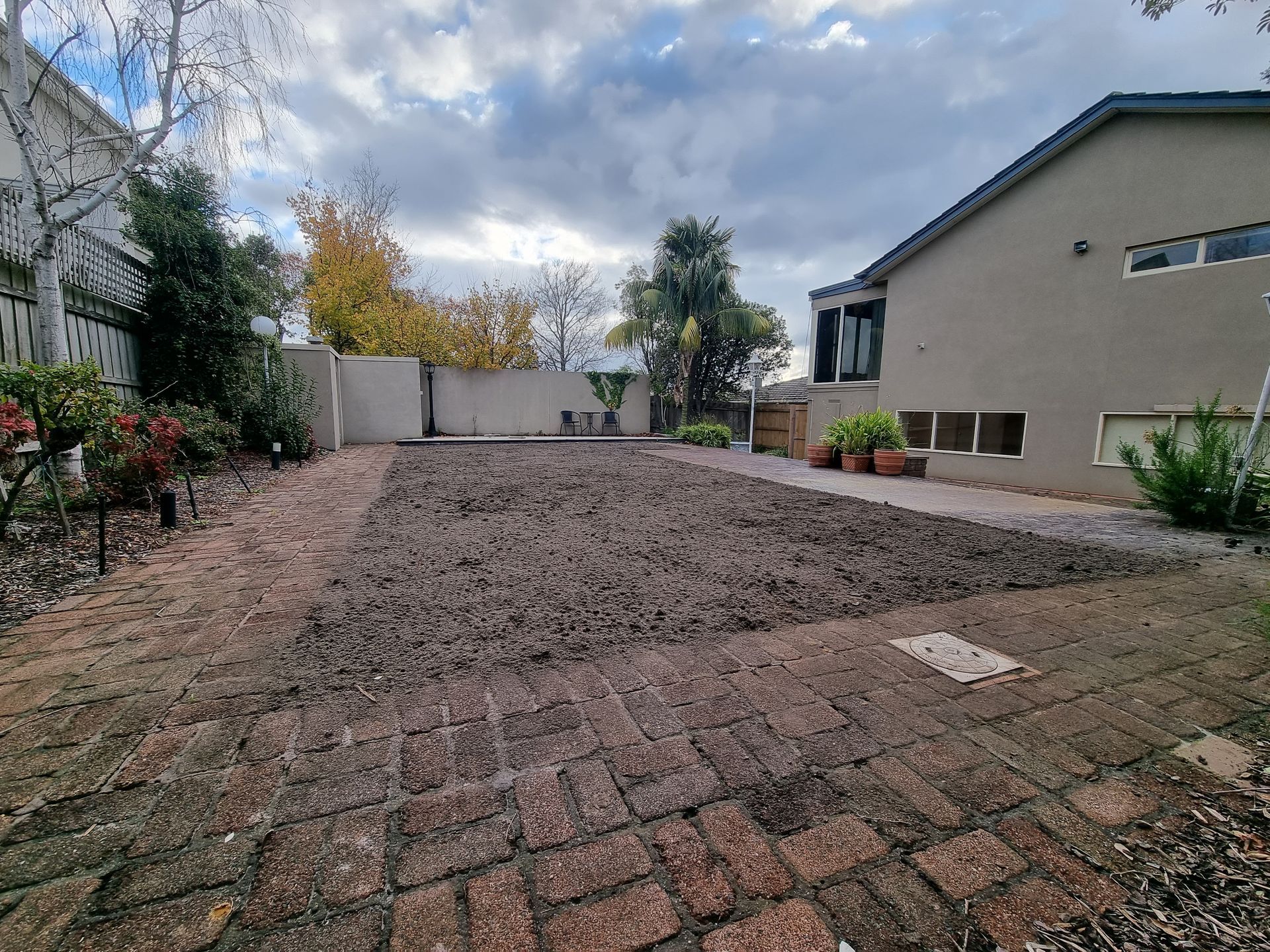 A dirt-covered yard beside a house, viewed from a brick patio under a cloudy sky — All Over Pool Removals in Doncaster, VIC