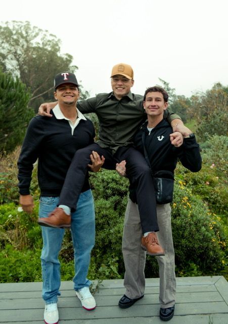 Three people outside; one person is being held up by the other two. They're on a deck with foliage behind them.