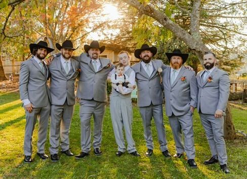 Groom and groomsmen in gray suits and cowboy hats pose outside with a baby.