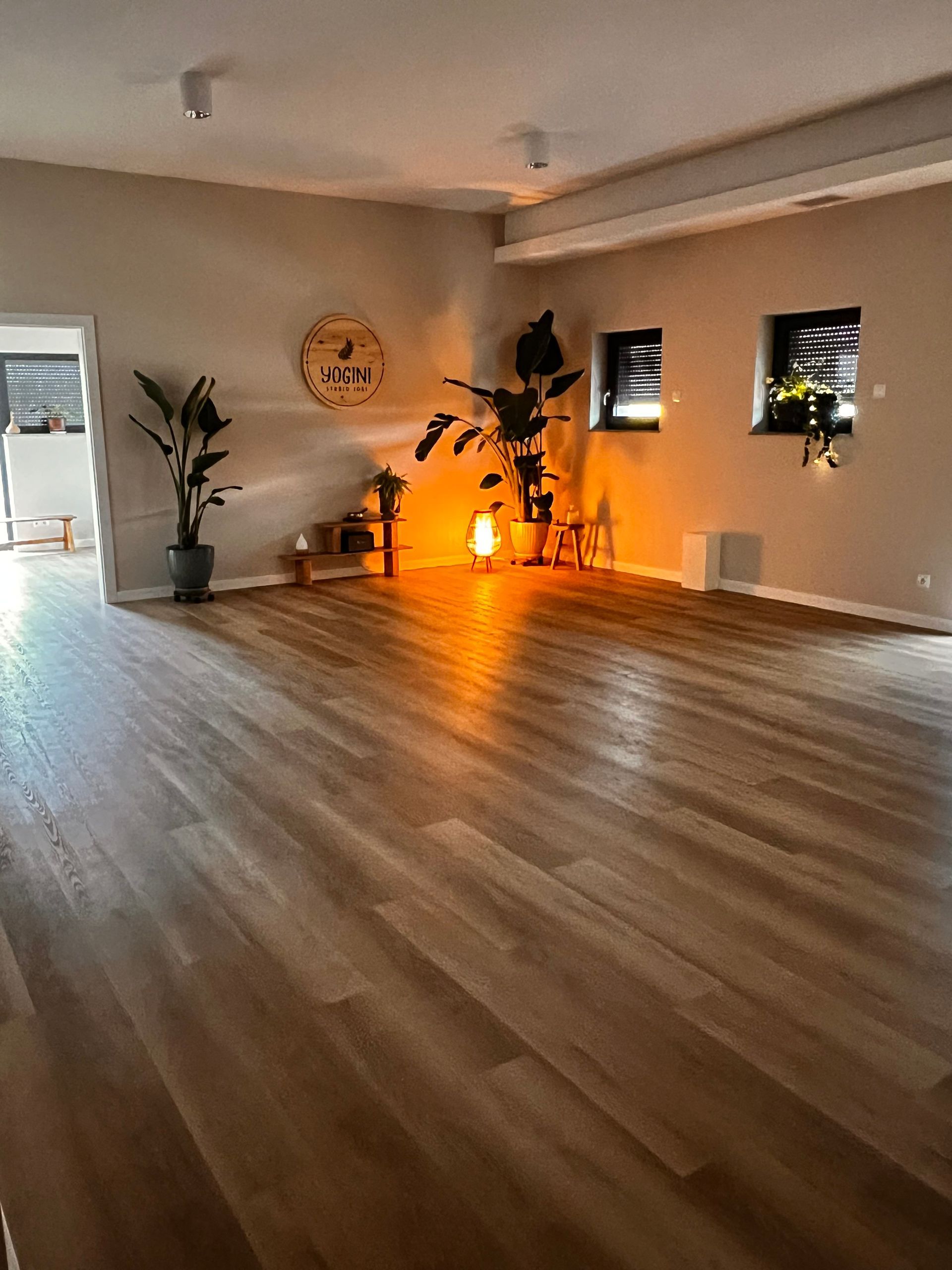 Spokojne studio jogi z drewnianą podłogą, dwiema dużymi roślinami doniczkowymi, świecącą lampą solną i ciepłym oświetleniem.