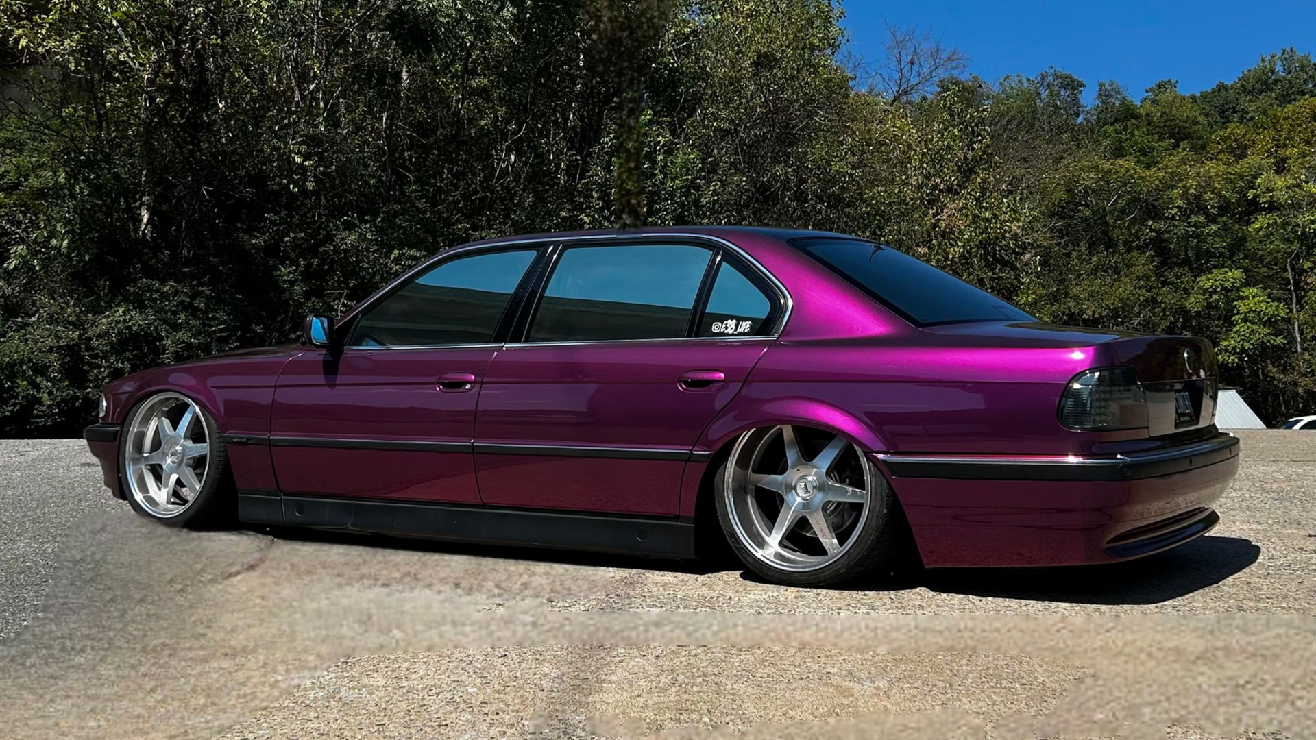 A low-slung, metallic purple sedan with large, polished silver wheels parked on a gravel surface against green trees.