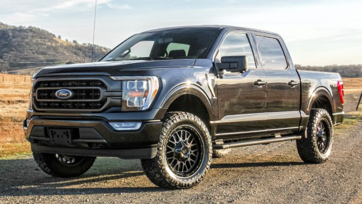 A dark gray Ford F-150 crew cab pickup truck with aftermarket off-road wheels, parked on a gravel path in a grassy field.
