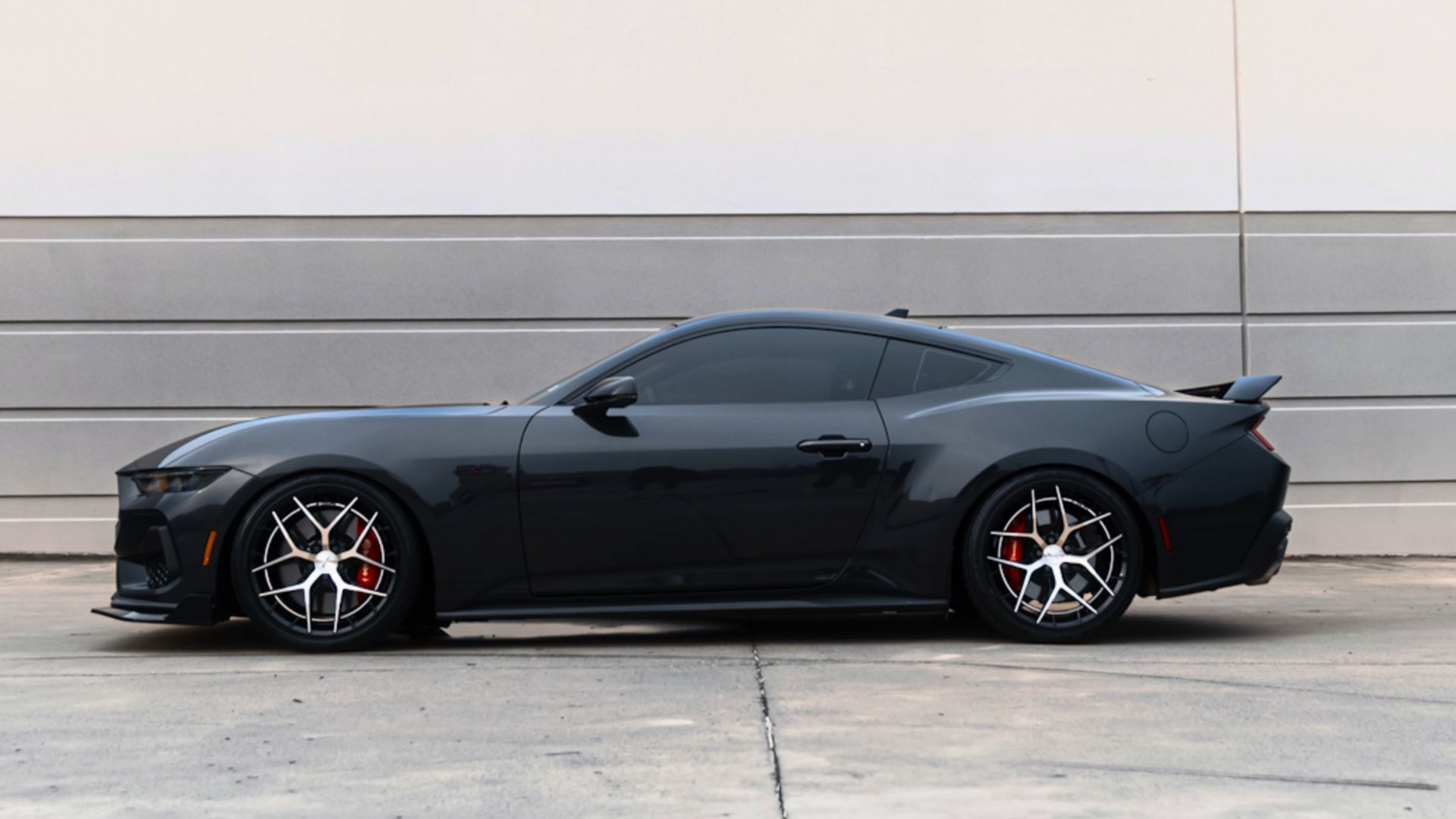 A black Ford Mustang parked in profile against a light gray wall, featuring dark tinted windows and custom silver wheels.