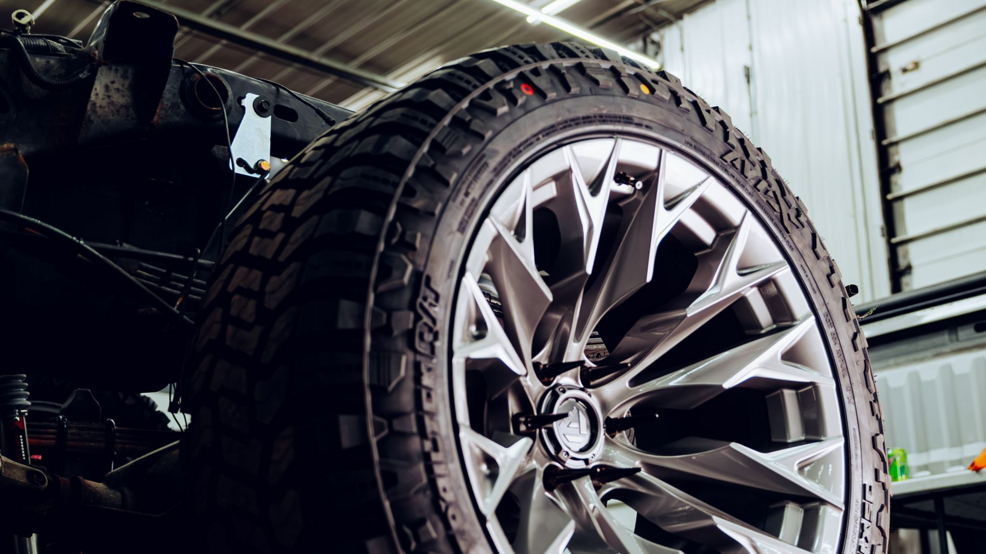 A close-up of a modern, angular silver alloy wheel with an off-road tire, installed on a vehicle inside a garage.