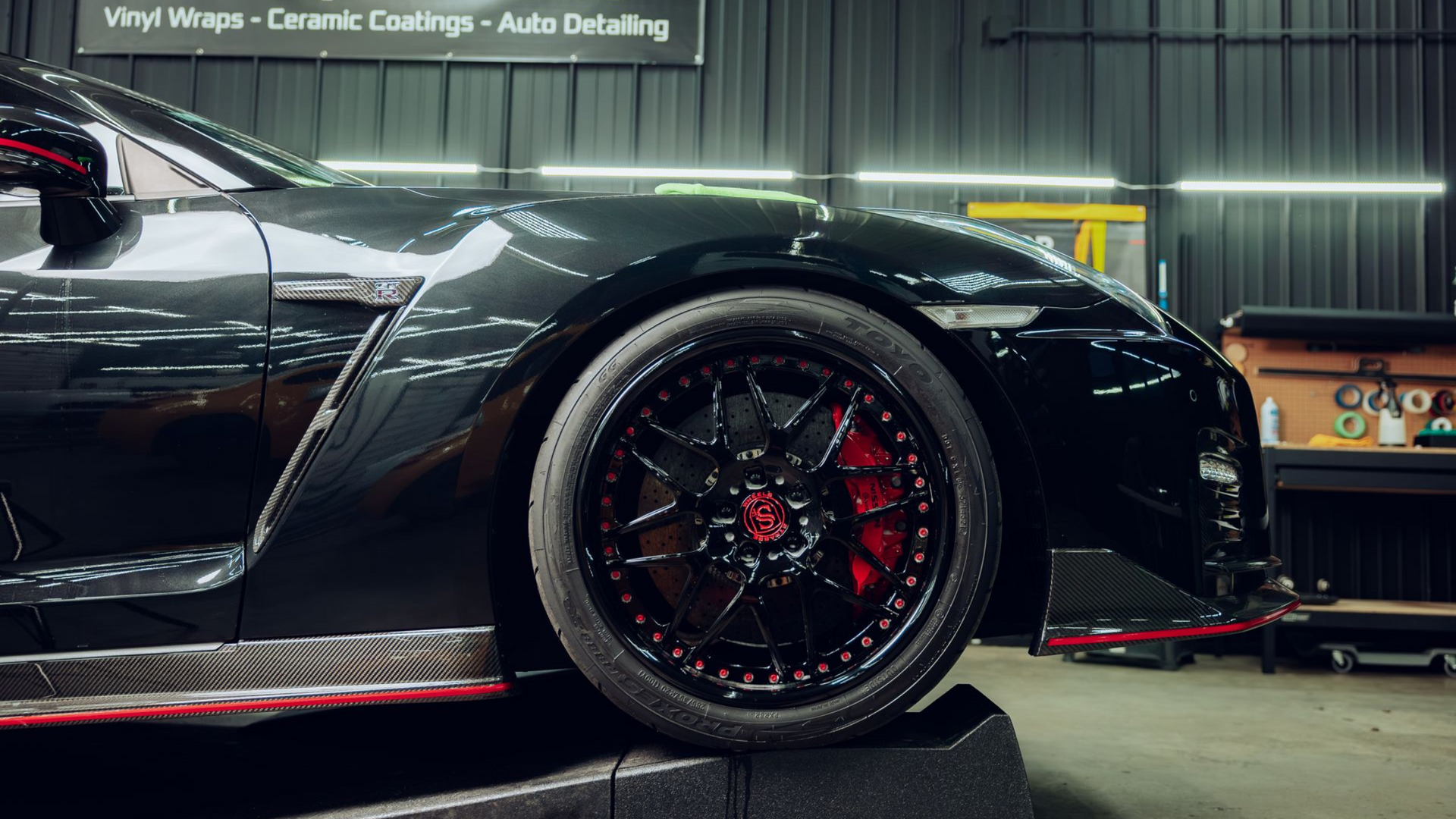 A black Nissan GT-R on a service ramp in a workshop, featuring red brake calipers and black rims with red accents.