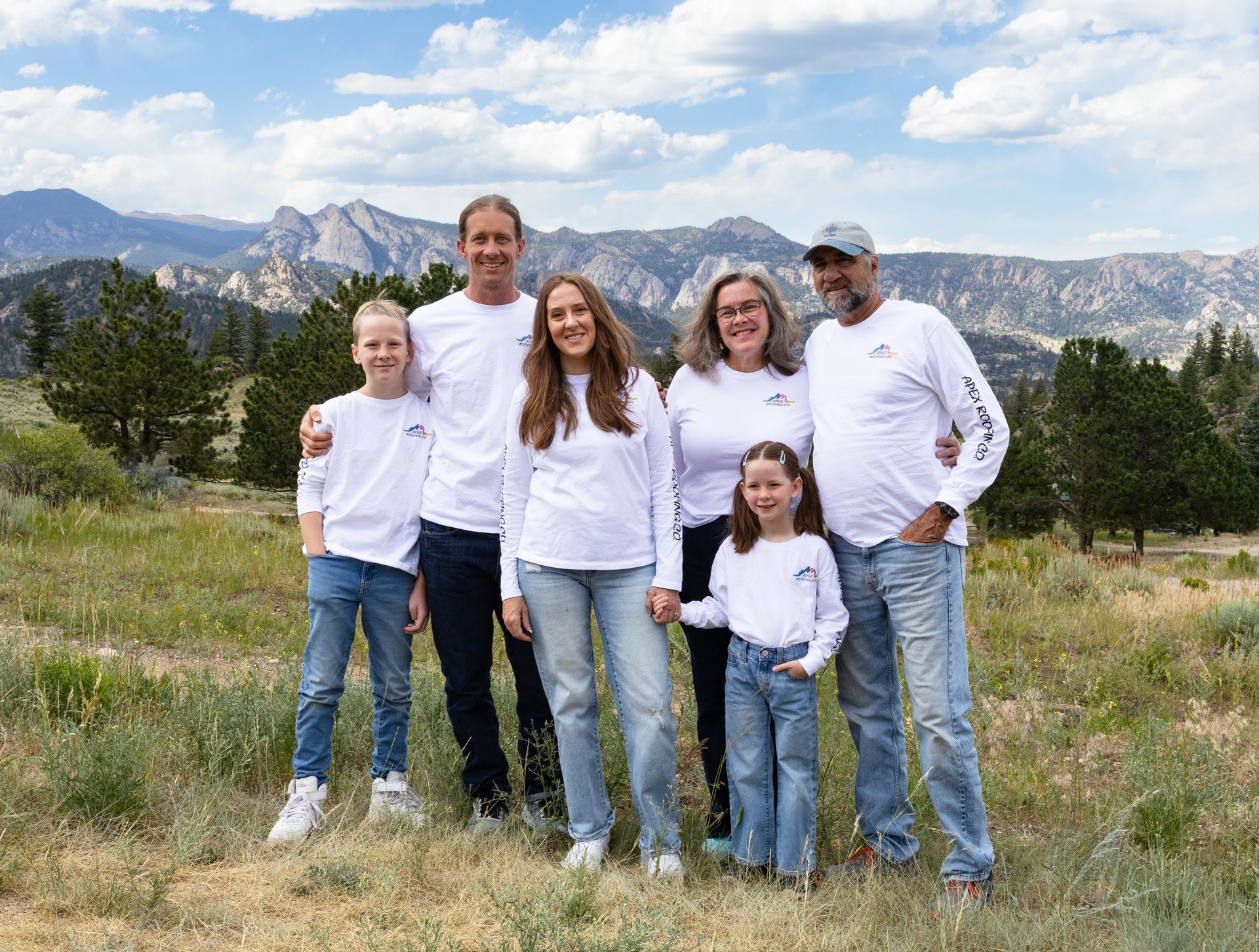 Family of six standing in a grassy field with mountains and trees in the background, wearing white tops.
