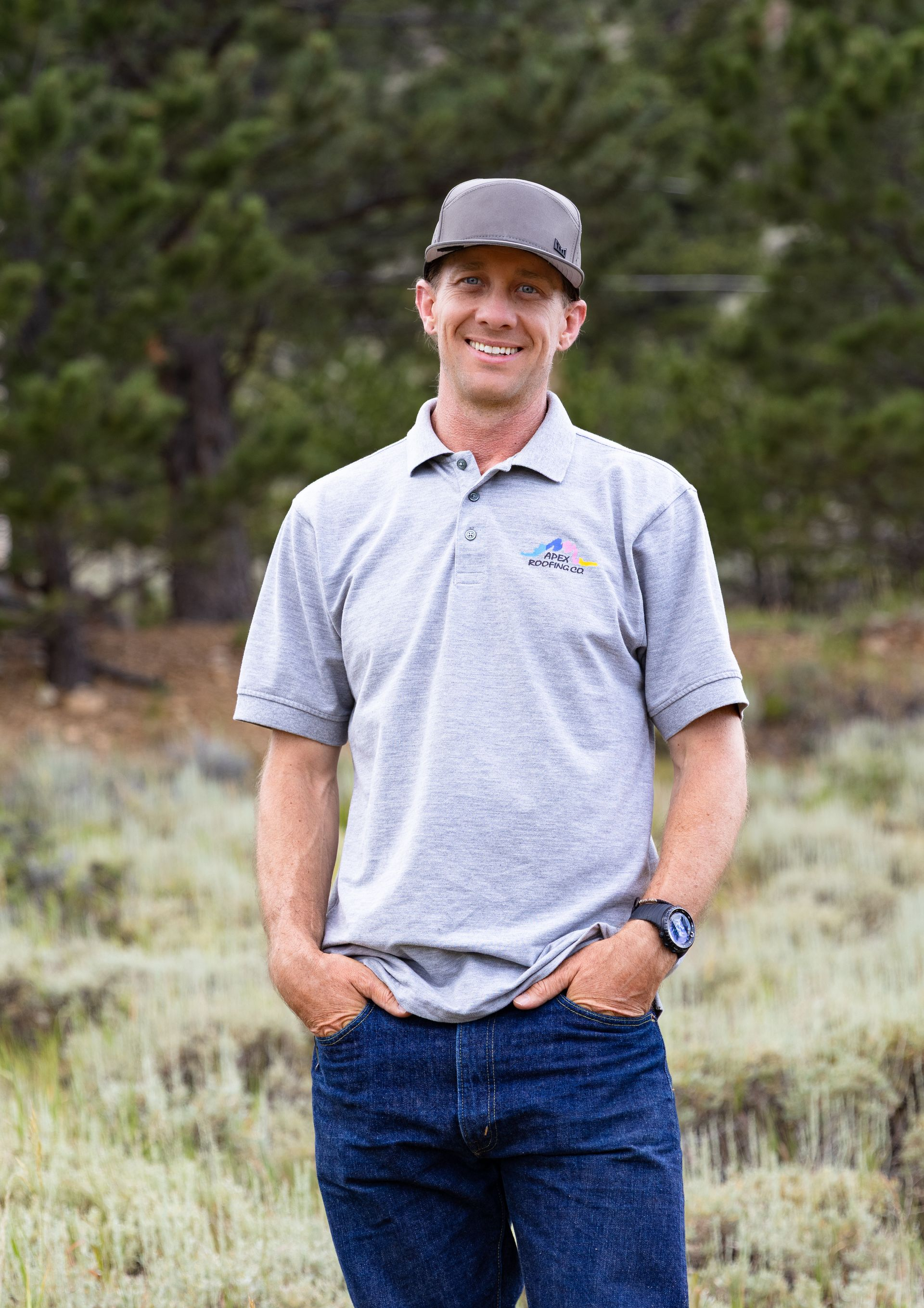 Man standing outdoors in a gray polo, jeans, and black cap, with hands in pockets and a slight smile.