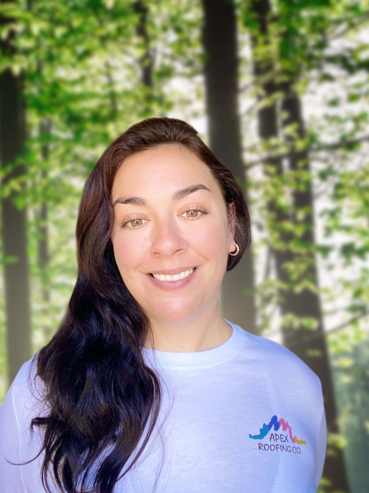 Smiling person in a white shirt with a mountain logo, standing before a blurred forest background