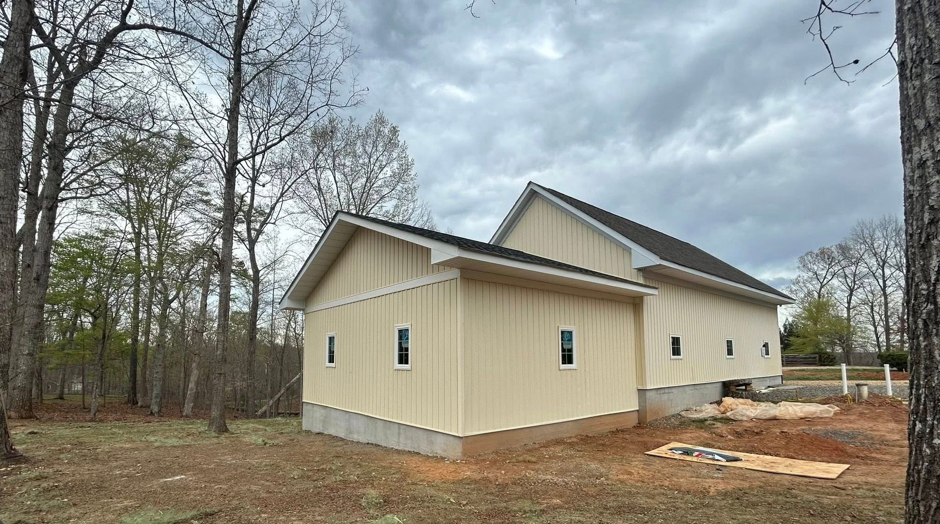 Single-story beige house under construction in a wooded area, with a cloudy sky and dirt foreground.