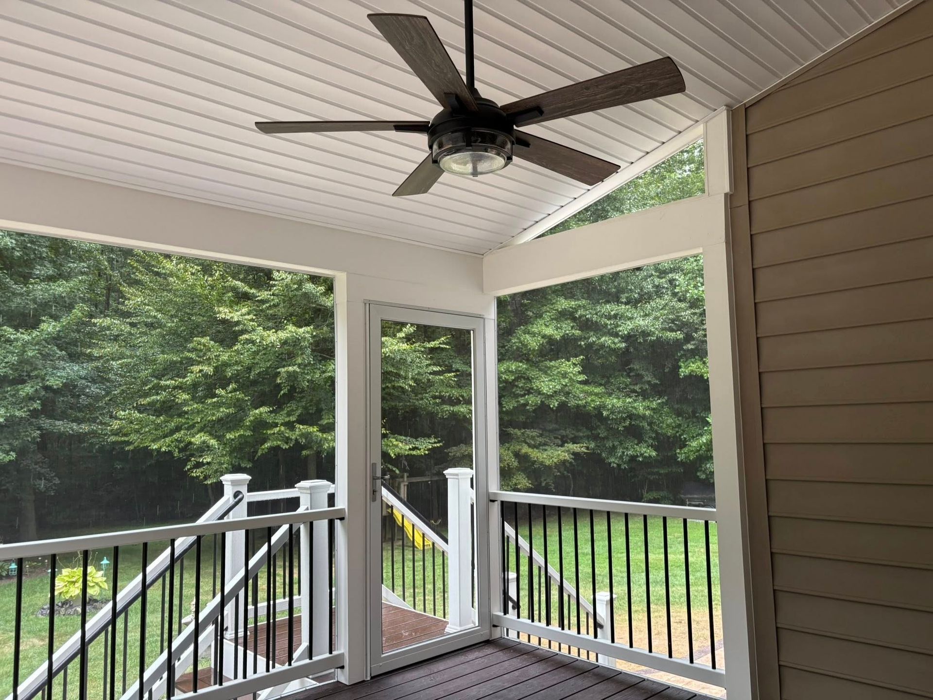 Covered porch with ceiling fan, white railings, and screened view of green trees.