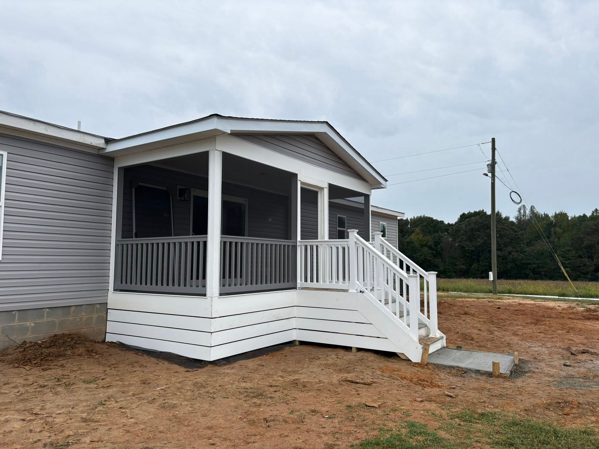 White porch-style modular home with front steps on a dirt lot under a cloudy sky