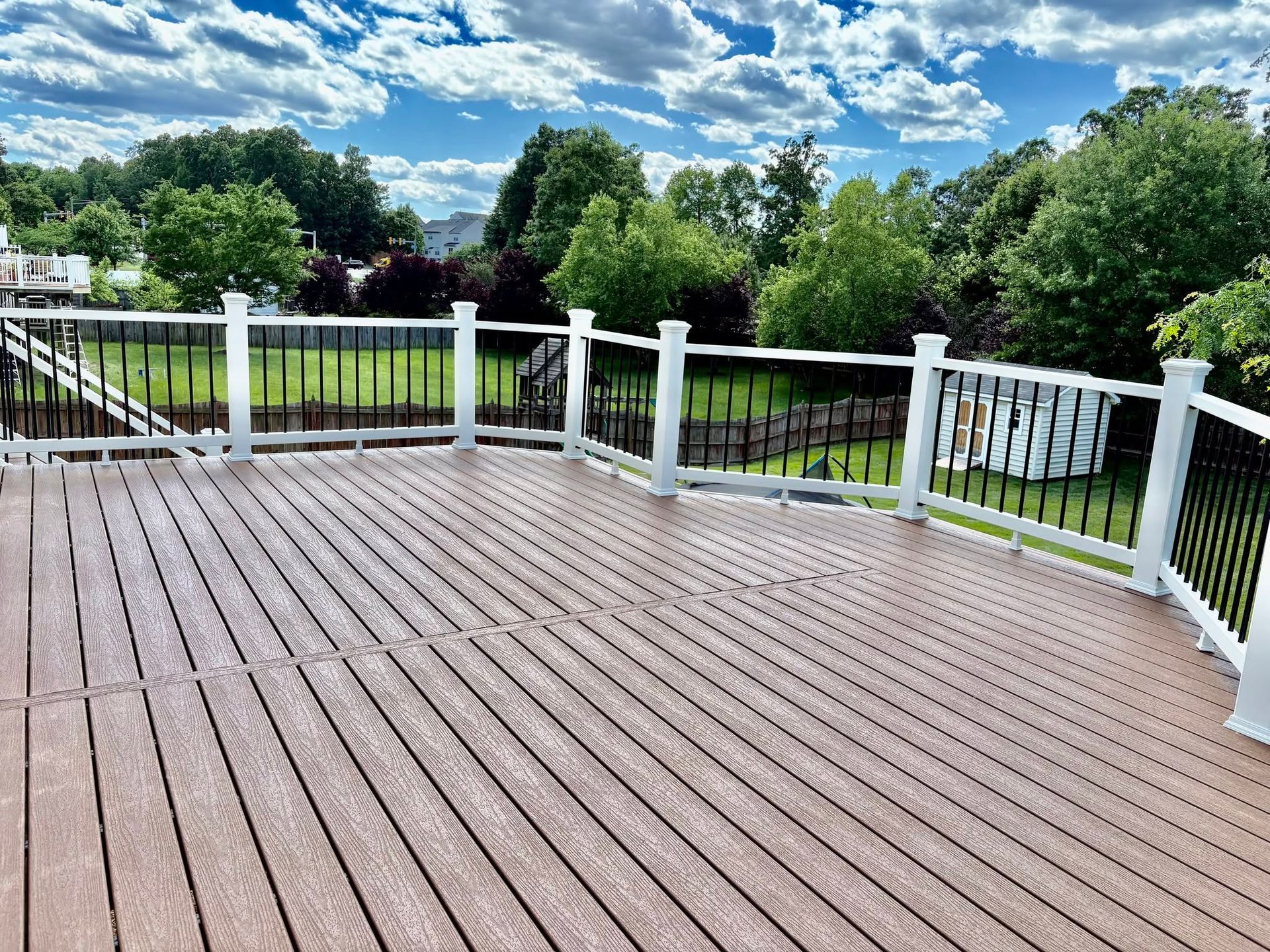 Wooden deck with white railing overlooking a green yard and trees under a partly cloudy sky