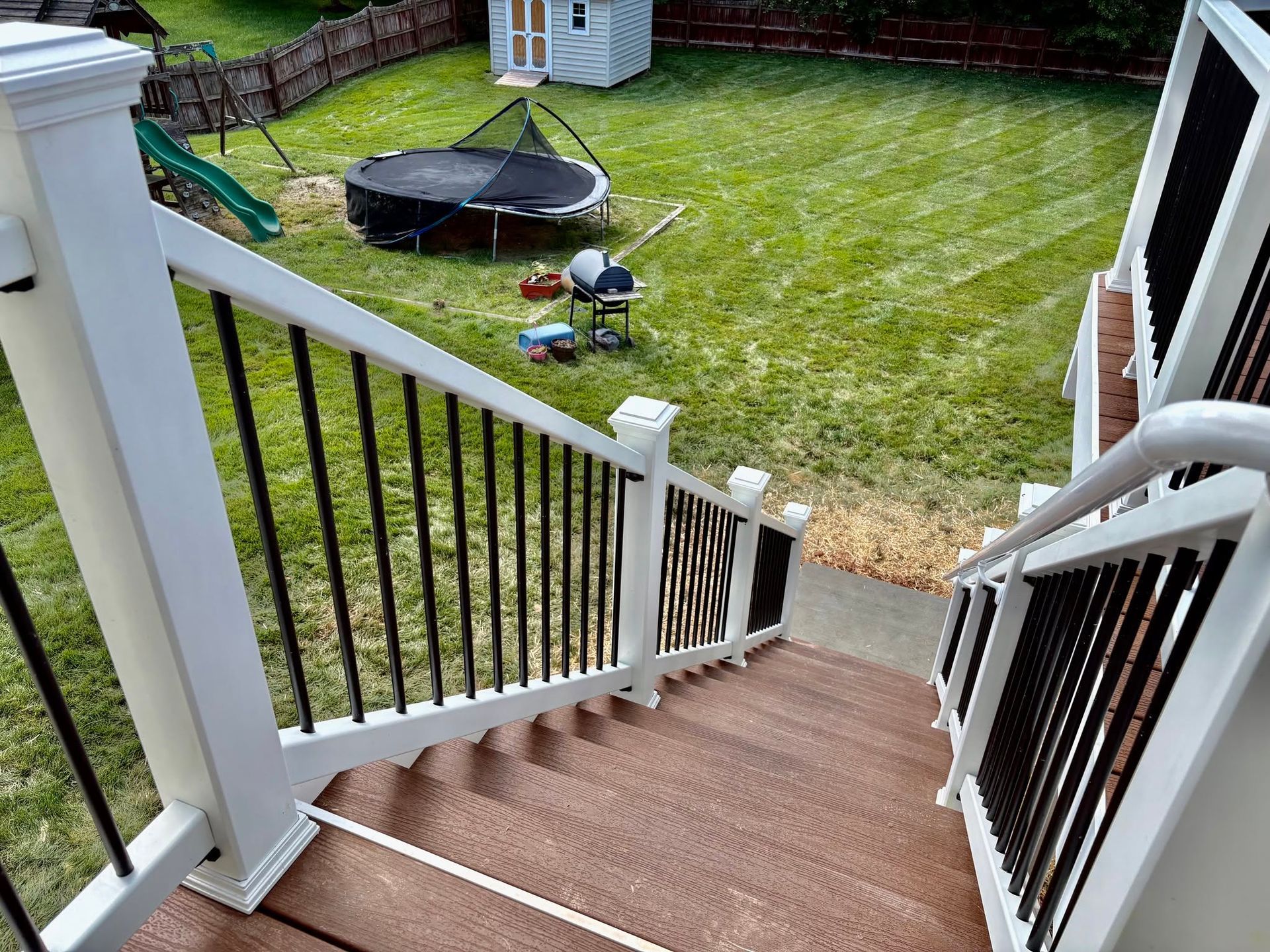 View from porch steps to a fenced backyard with a trampoline, play toys, and green lawn.