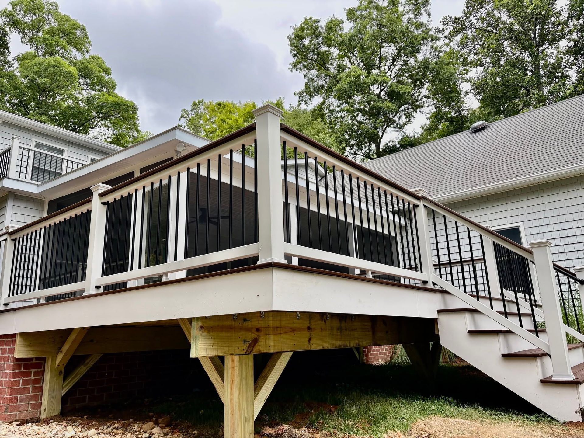Raised wooden deck with white railings and stairs beside a house, surrounded by trees
