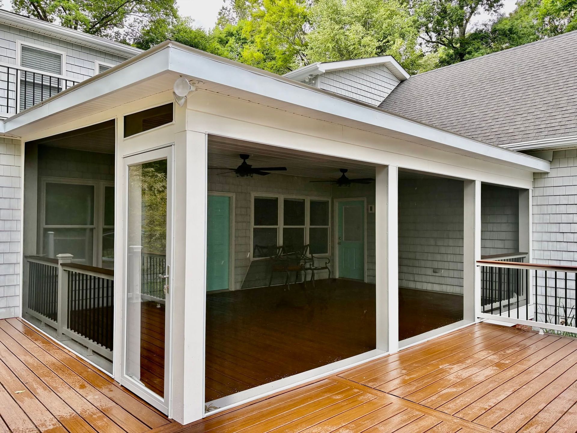 Screened porch with white trim and a wooden deck beside a house, with green trees overhead
