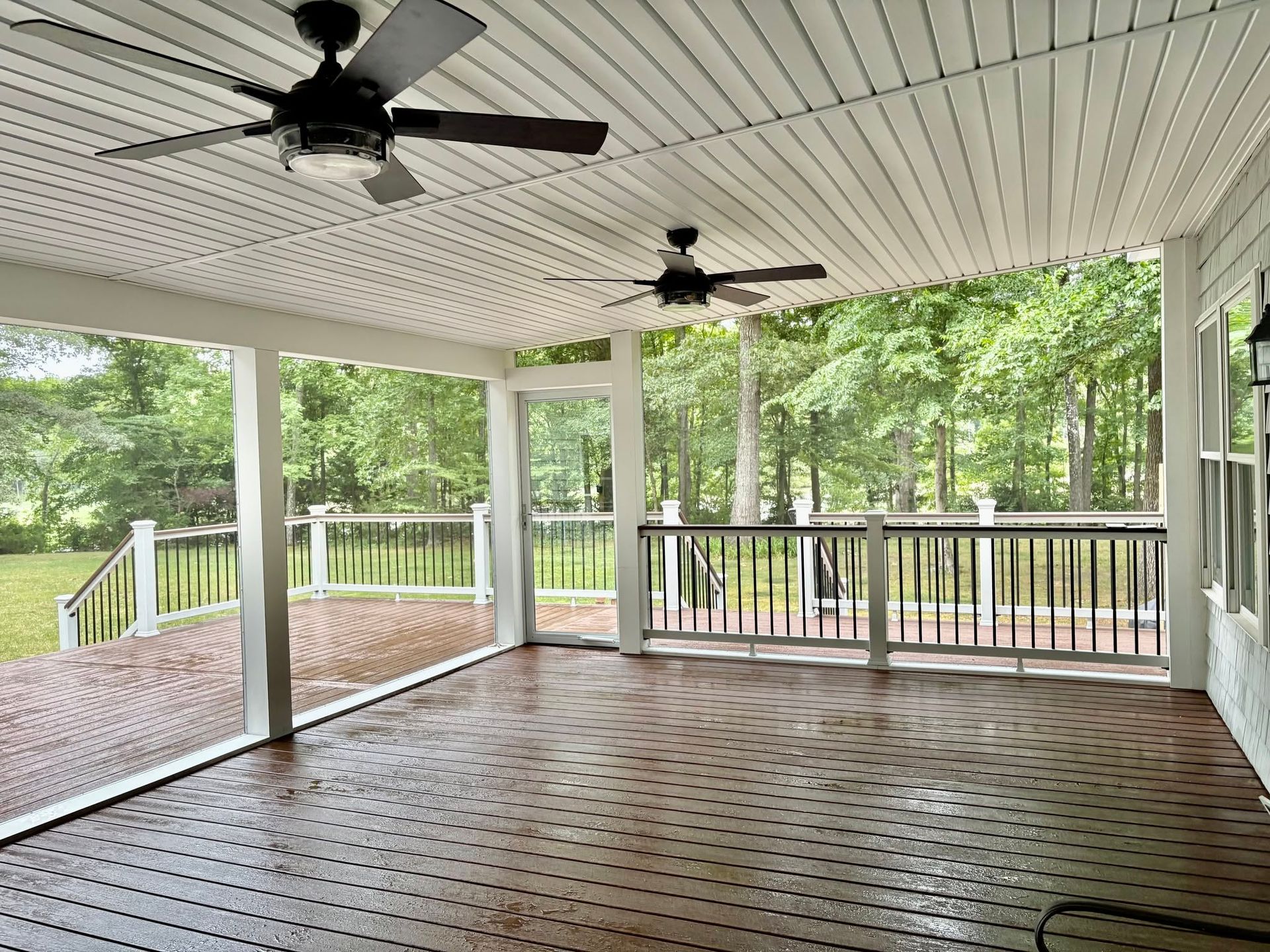 Covered wooden porch with white ceiling, black ceiling fans, and railing overlooking a green wooded yard