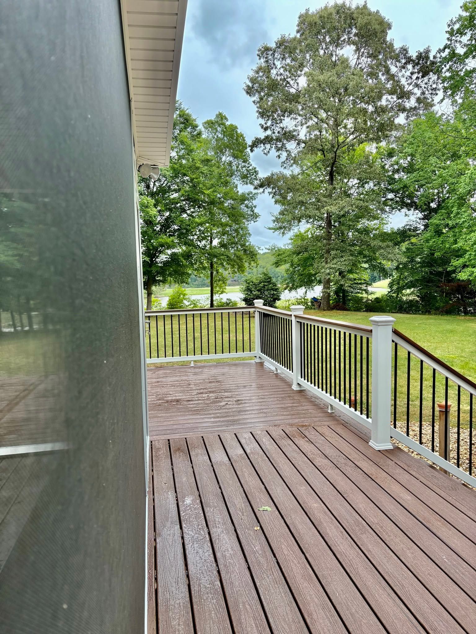 Wooden deck with white railing beside a house, overlooking a green yard and trees.