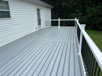 Gray wooden deck attached to a white house, with a railing along one side and trees beyond.