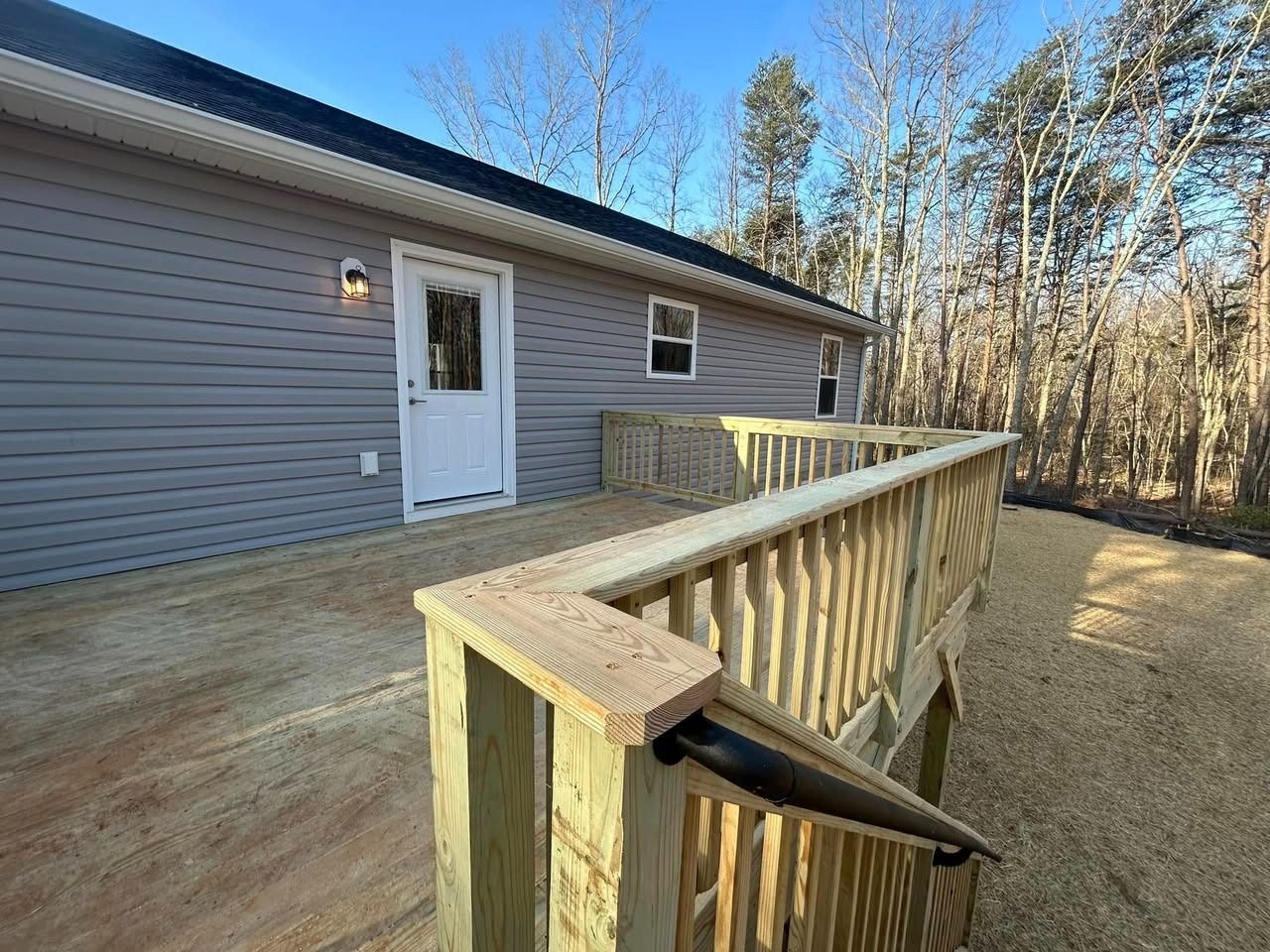 Wooden ramp with railing leading to a white door on a house exterior, beside a gravel yard and trees