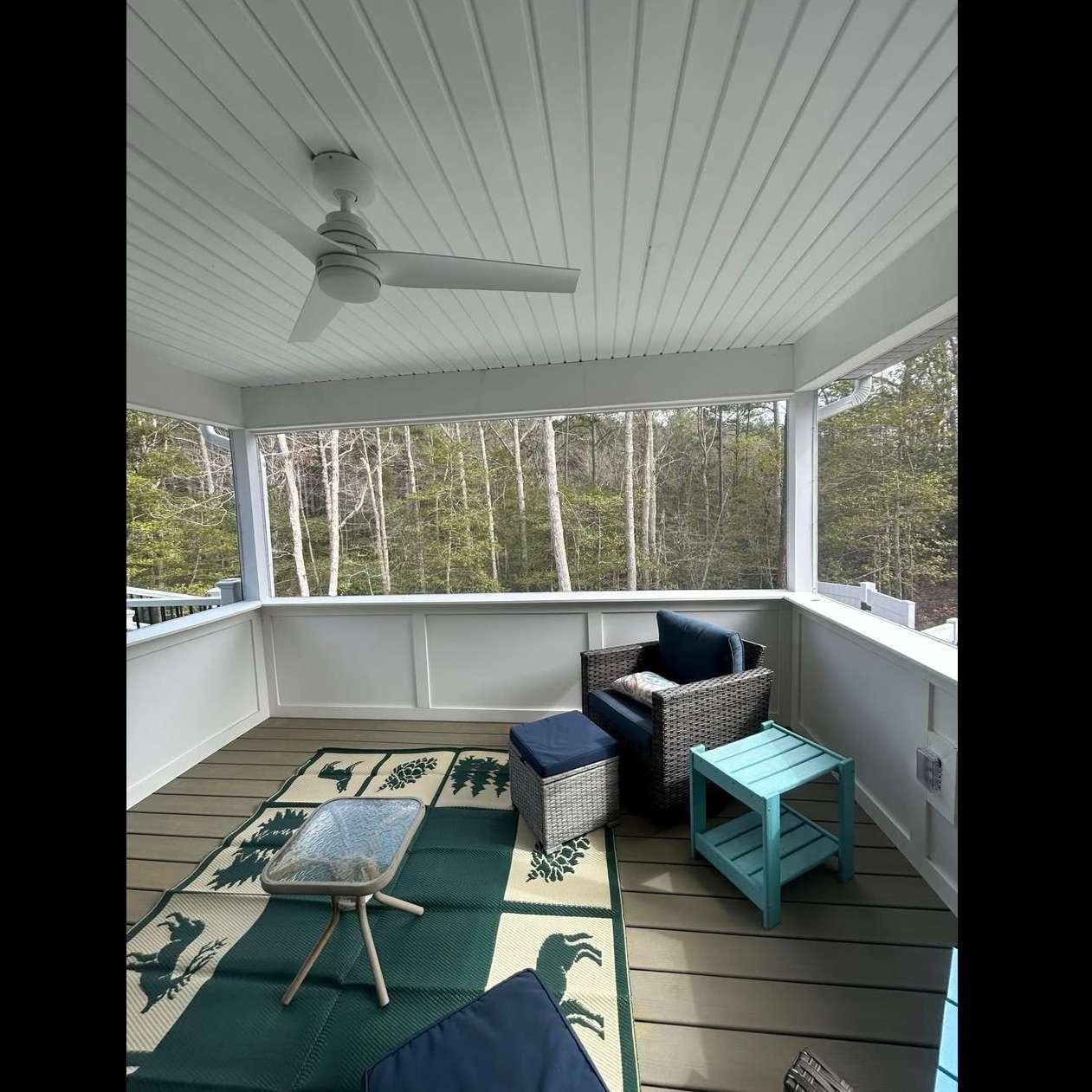 Covered porch with white ceiling fan, blue seating, and a green rug overlooking trees