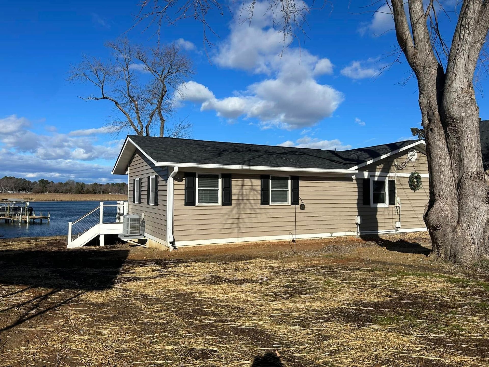 Lakeside beige house with white trim, dock, and leafless trees under a bright blue sky