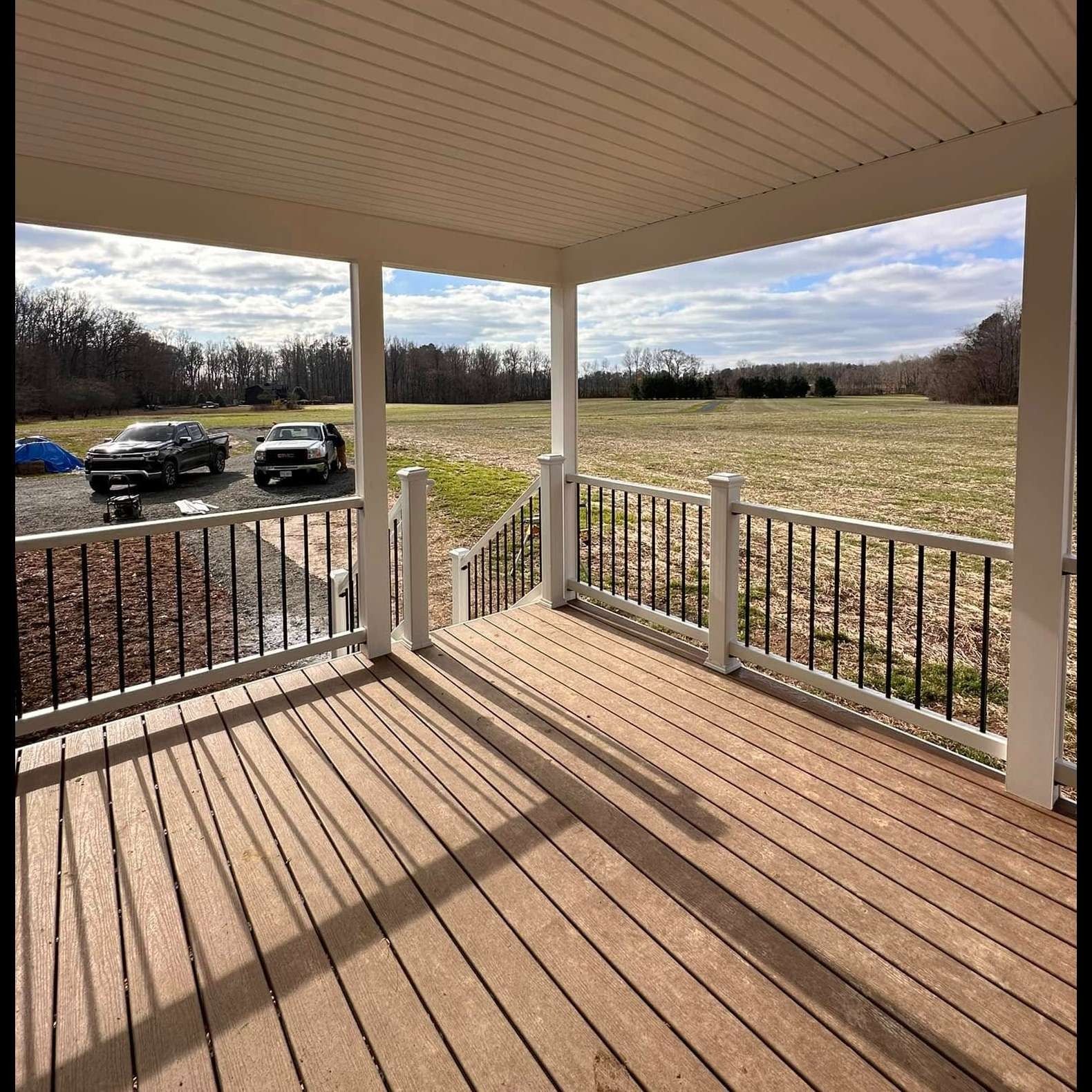 Covered porch with white railings overlooking a grassy field and parked cars in the distance.