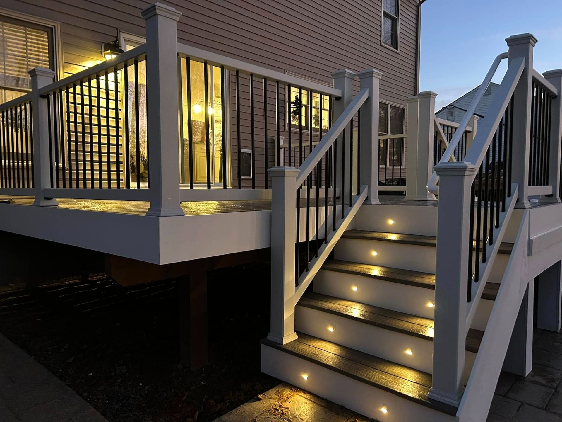 Lit wooden stairs with railings leading to a raised deck beside a brick house at dusk.