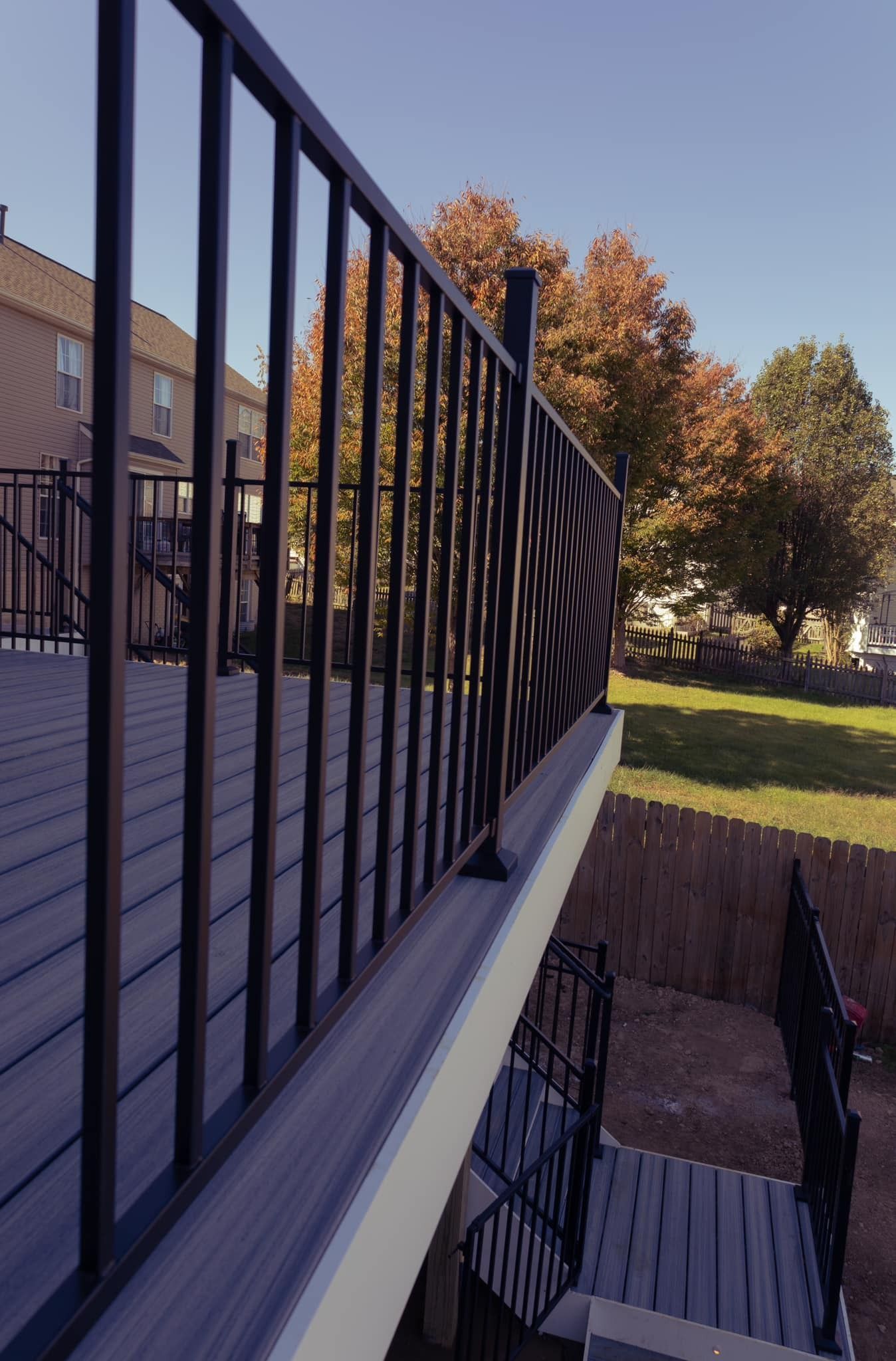 Black metal railing beside a wooden staircase overlooking a sunlit backyard with trees and a house