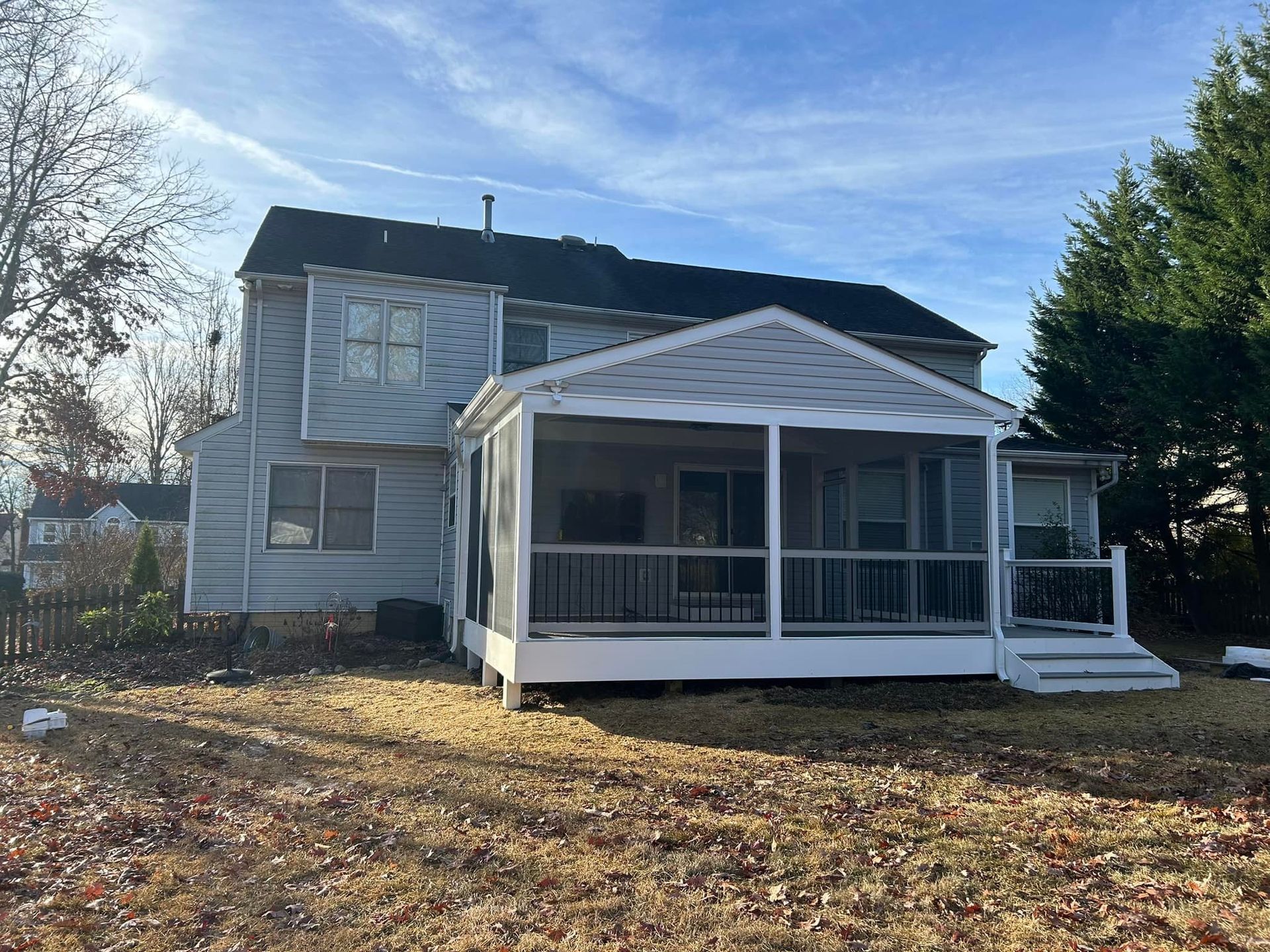 Two-story gray house with a white screened porch and steps, surrounded by fallen leaves under a blue sky